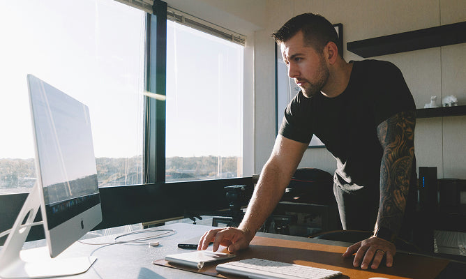 A tattooed man standing at a desk working on his Apple computer.